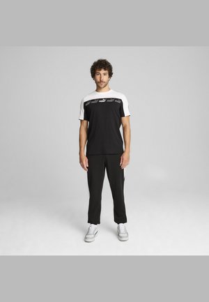 Svart t-shirt med vita ärmar, med en Puma-logotyp designad över bröstet. Kombinerad med svarta byxor och vita träningsskor.