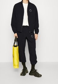 Marineblauwe trackjack met rits en logo, gecombineerd met bijpassende joggers. Zwarte sneakers en een gele schoudertas maken de look compleet.