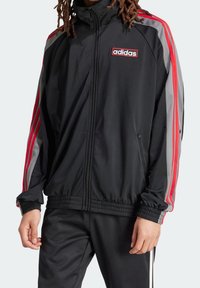 Chaqueta atlética negra con cremallera y rayas rojas y grises a lo largo de los hombros. Presenta puños y cintura elásticos, con un logo de Adidas en el pecho.