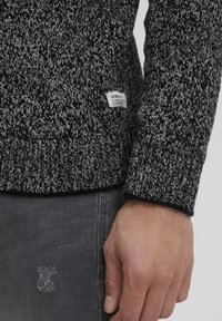 Schwarz-weißer Strickpullover mit einem strukturierten Muster, ausgestattet mit einem gerippten Saum und Bündchen. Kleines Markenlabel an der unteren Seite.