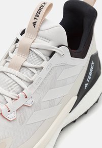 Lys grå treningssko med mesh-overdel, kontrasterende svart hæl, beige detaljer og rød søm. Har en solid såle og fremtredende merkevare.
