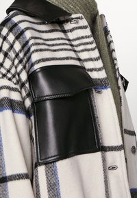 Plaid shirt jacket in cream, black, and blue tones. Features a black leather pocket and a dark collar. Soft wool blend texture.