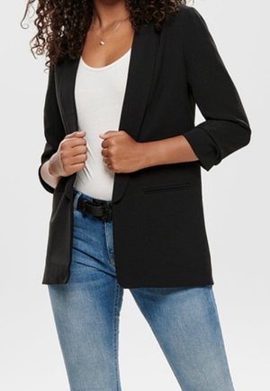 Mujer con cabello rizado que lleva una blazer negra sobre una camiseta blanca y vaqueros azules, sujetando las solapas de la blazer con ambas manos.