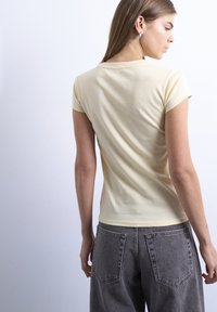 Γυναίκα με μακριά καστανά μαλλιά, φορεμένη με ανοιχτό κίτρινο κοντομάνικο πουκάμισο και σκούρα γκρι τζιν, με την πλάτη προς την κάμερα σε φωτεινό φόντο.