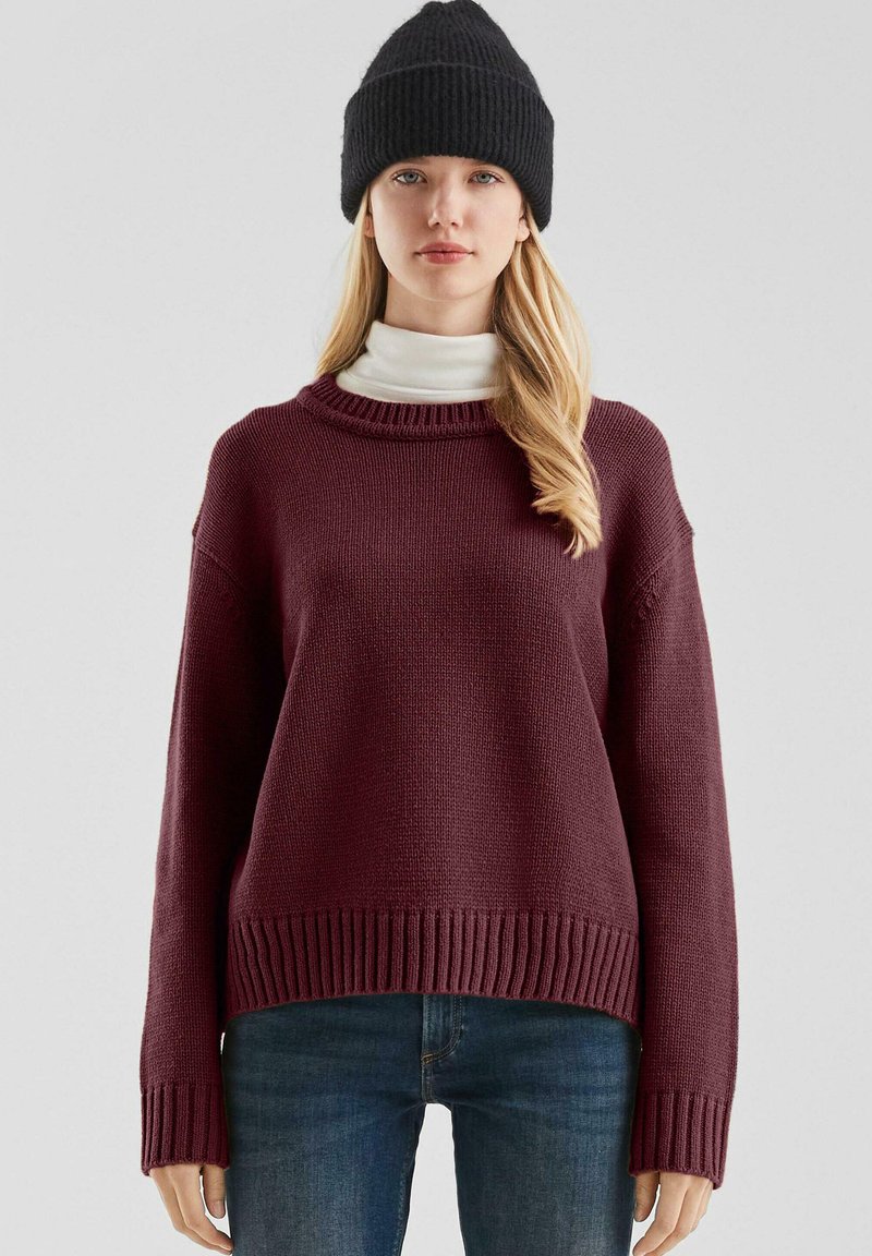 Bordeaux gebreide trui met een geribde halslijn en zoom, met verlaagde schouders en lange mouwen, laag over een witte turtleneck.