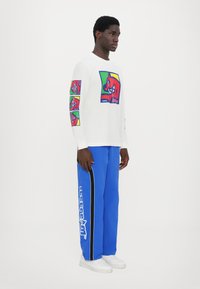 Weißes Longsleeve-Shirt mit bunten Grafikdrucken auf der Vorderseite und den Ärmeln. Helle blaue Hose mit weißem Logo und schwarzem Seitenstreifen. Weiße Sneaker.