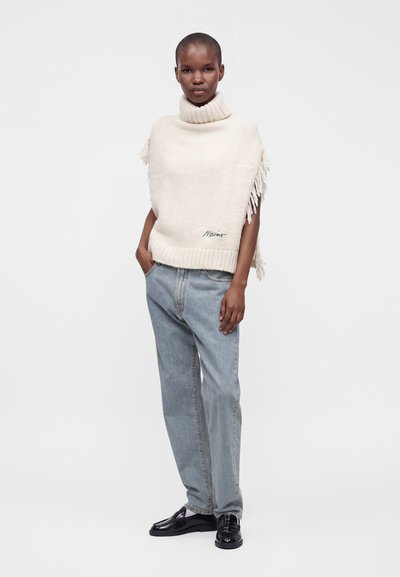Mannequin portant un pull sans manches à col roulé crème avec des franges, un jean bleu clair et des mocassins noirs, debout avec une main dans la poche.