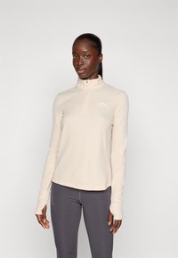 Pull long à manches longues beige clair avec un col zippé, en tissu texturé, et un logo blanc sur la poitrine gauche, associé à des leggings gris foncé.