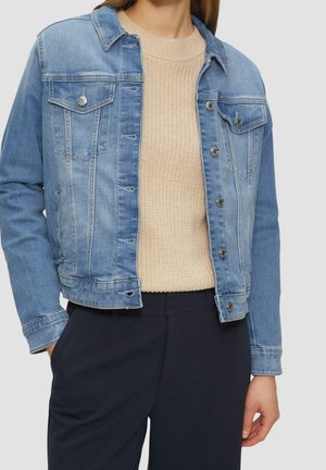 Vrouw draagt een blauwe spijkerjas over een beige gebreide trui en donkere broek, staand tegen een lichte achtergrond.