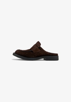 Zapatilla estilo mule de textura de piel marrón con parte superior suave y tira lateral. Presenta una suela plana negra con una ligera curva y sin adornos visibles.