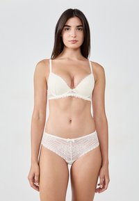 Kremowy koronkowy biustonosz z falbankami i regulowanymi ramiączkami, w połączeniu z pasującymi majtkami koronkowymi z małym kokardkowym detalem z przodu.