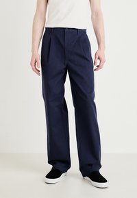 Pantalon bleu marine à coupe décontractée, doté de plis à l'avant et d'une fermeture à bouton. Tissu en mélange de coton. Assorti à des baskets noires.