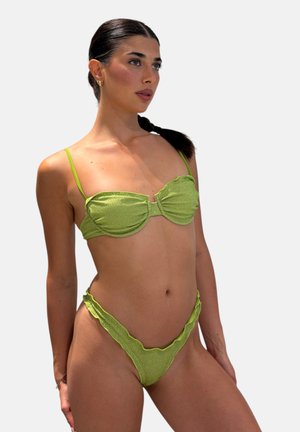 Donna con capelli scuri raccolti in una coda bassa che indossa un bikini verde chiaro strutturato con slip a vita alta con volant su sfondo semplice.