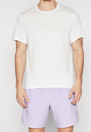 Man wearing white short-sleeve Nike sports shirt and light purple Nike shorts, standing against plain background.