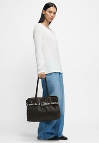 Brązowa skórzana torba typu tote o wyraźnej formie, z ozdobną srebrną klamrą i detalami na pasku. Modelka trzyma ją w codziennym stroju.