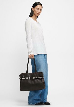 Brązowa skórzana torba typu tote o wyraźnej formie, z ozdobną srebrną klamrą i detalami na pasku. Modelka trzyma ją w codziennym stroju.