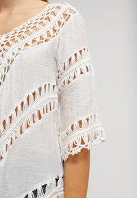 Blouse blanche texturée avec des motifs de crochet ouverts sur les manches, le col et le corps, portée par une personne avec l’épaule et le haut du bras découverts.