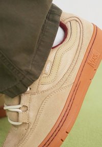 Adidaș bej din ante cu un panou lateral din plasă, șireturi albe, talpă din cauciuc portocalie și accente subtile de vișiniu. Prim-plan al pantofului pe un fundal verde.