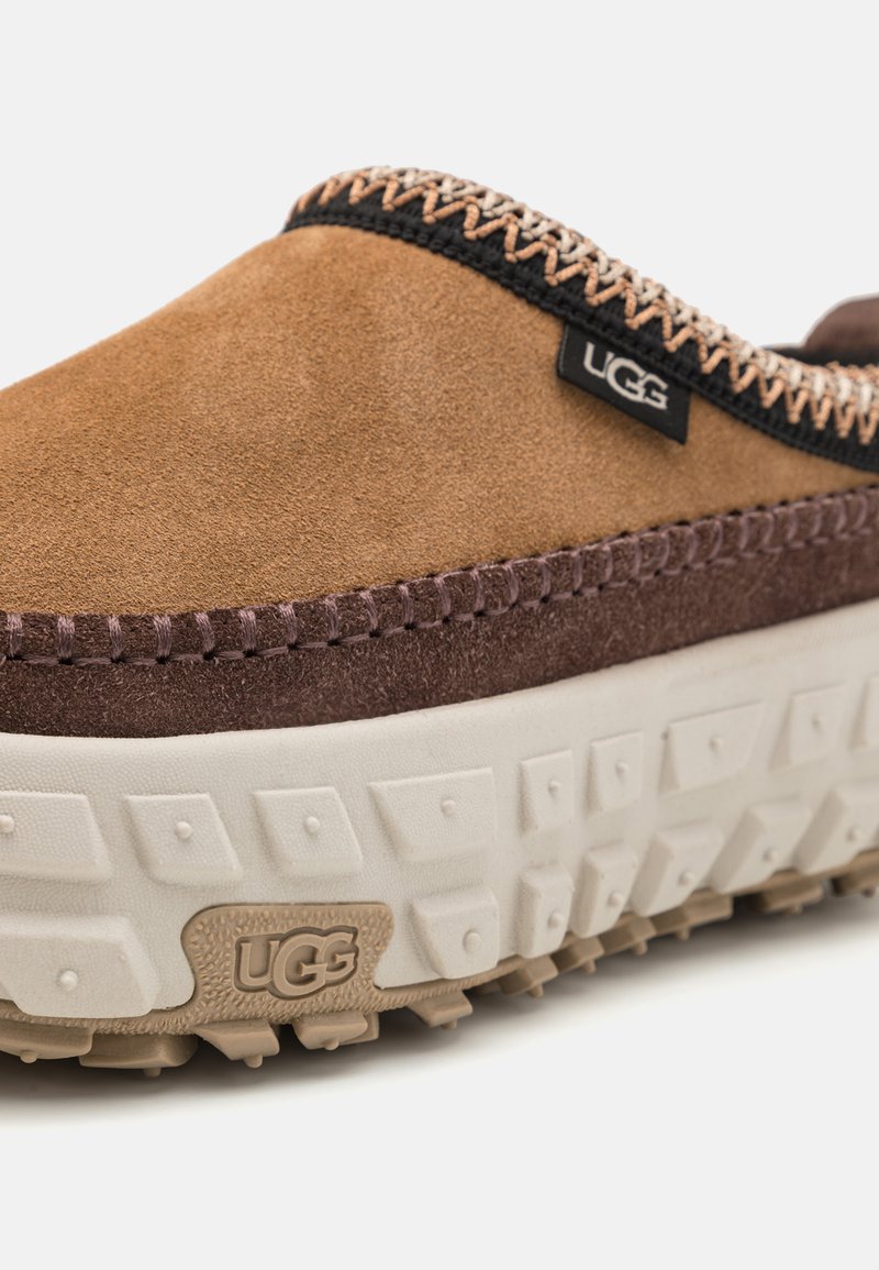 Zapato slip-on de ante marrón con un acento texturizado marrón, suela de goma crema y costuras en contraste. Presenta el logo de UGG en el lateral.
