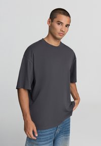 Ciemnoszara, oversize'owa koszulka wykonana z miękkiej bawełny, z krótkim rękawem, okrągłym dekoltem i luźnym krojem. W połączeniu z jasnoniebieskimi dżinsami.