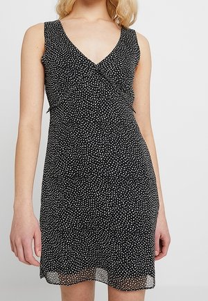 Vestido negro sin mangas con lunares blancos, escote en V, cintura ajustada y dobladillo a la altura de la rodilla, llevado por una persona de piel clara y cabello rubio.