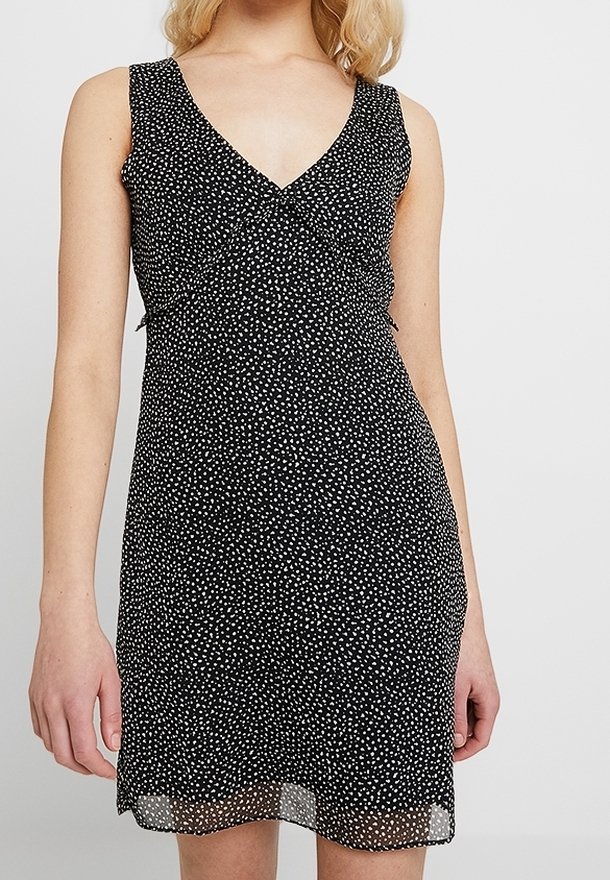 Robe noire sans manches à pois blancs, col en V, taille cintrée et ourlet longueur genou portée par une personne à la peau claire et aux cheveux blonds.