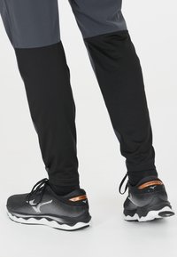 Schwarze und graue Sporthose mit tailliertem Design, aus strukturiertem Material und mit gerippten Bündchen, kombiniert mit schwarzen Laufschuhen mit weißen Sohlen.