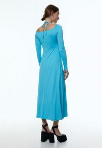 Langärmliges, figurbetontes Kleid aus glattem blauen Stoff mit quadratischem Ausschnitt, offenem Rücken und Bindeschluss. Kombiniert mit schwarzen Plateauschuhen.
