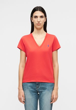 Γυναίκα με μακριά σκούρα μαλλιά, ντυμένη με κόκκινη μπλούζα V λαιμόκοψη και μπλε τζιν, στέκεται μπροστά σε απλό ανοιχτό γκρι φόντο.