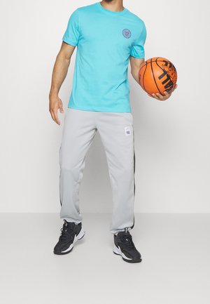 Homme en t-shirt turquoise, pantalon de survêtement gris et baskets noires tenant un ballon de basket orange sur un fond uni.