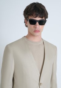 Blazer beige su misura con rever a collo a lancia, un bottone e texture morbida, abbinate a una maglietta a collo rotondo color beige chiaro e occhiali da sole neri.