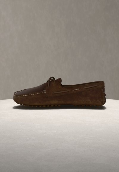 Καφέ δερμάτινο μοκασίνι με ραμμένες λεπτομέρειες και λαστιχένια σόλα με μοτίβο από πέτρες, εμφανιζόμενο από το πλάι σε ουδέτερο υπόστρωμα και φόντο.