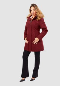 Cappotto borgogna con cappuccio foderato in pelliccia, design aderente, maniche lunghe, tasche laterali e chiusura con zip, abbinato a pantaloni neri a zampa d'elefante e tacchi.