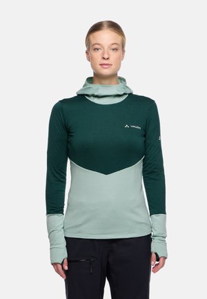 Femme portant un haut d'extérieur à manches longues vert et bleu clair avec des ouvertures pour les pouces et une capuche, se tenant contre un fond blanc uni.