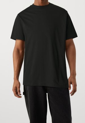 Mand iført en ensfarvet sort t-shirt med korte ærmer og rund hals og mørke bukser mod en neutral baggrund.
