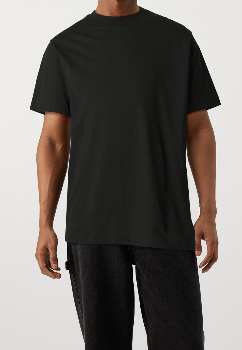 Homme portant un t-shirt noir uni à manches courtes et col rond, avec un pantalon sombre sur un fond neutre.