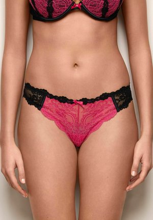 Mujer con sujetador de encaje rosa y negro y ropa interior a juego, de pie frente a un fondo neutro.