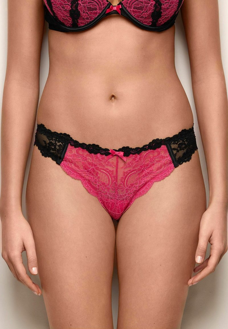 Frau trägt rosa-schwarzen Spitzen-BH und passende Spitzen-Unterwäsche, steht vor einem neutralen Hintergrund.