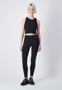Zwart sportcrop top en strakke leggings met zijzakken. Model draagt zwarte sportschoenen met rode en blauwe accenten. Glad textuur.