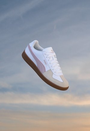 Baskets blanches avec une bande rose clair, embout beige et semelle marron, montrées en train de flotter sur un fond de ciel bleu et orange.