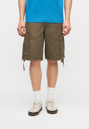 Άνδρας με καφέ cargo σορτς, λευκές κάλτσες, μπεζ αθλητικά παπούτσια και μπλε πουκάμισο, στέκεται με τα χέρια χαλαρά δίπλα στο σώμα.