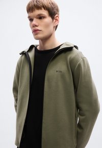 Χακί ζακέτα με φερμουάρ και κουκούλα, κατασκευασμένη από ένα ομαλό ύφασμα. Μαύρο λογότυπο στο στήθος, συνδυασμένο με μαύρο μπλουζάκι με στρογγυλή λαιμόκοψη από κάτω.