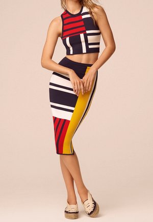 Vrouw draagt een mouwloze cropped top en een hoog getailleerde rok met rode, gele, witte en zwarte geometrische patronen, staand met gevouwen handen.