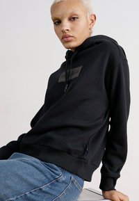 Svart hettegensar laget av mykt stoff med et brystlogo, med ribbede mansjetter og kant, stylet med lyseblå jeans.