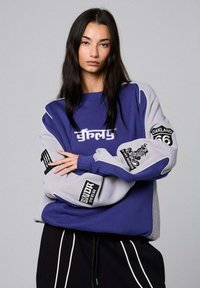 Felpa grande viola e grigia con grafiche e toppe di testo, polsini a coste e vestibilità rilassata. Pantaloni neri con righe bianche.