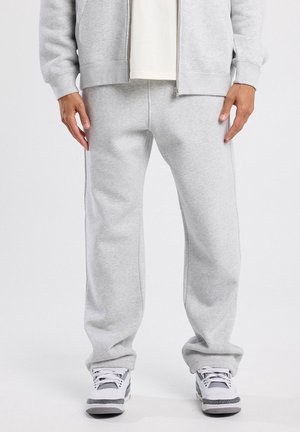 Homme portant un pantalon de survêtement gris clair, un sweat à capuche zippé gris, des baskets blanches, debout devant un fond uni clair.