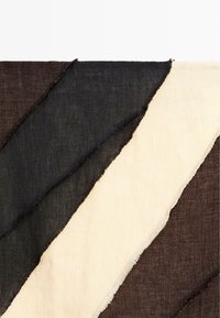 Diagonale Stoffstreifen in Schwarz, Beige und Dunkelbraun mit ausgefransten Kanten, überlappend auf weißem Hintergrund.