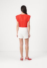 Haut perforé rouge avec des manches flottantes, short blanc taille haute, et mules à talons rouges. Texture lisse, coupe décontractée, et détails de design superposés.