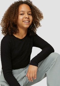 Zwarte, lange mouwen, textuurshirt met een ronde hals, gecombineerd met lichtgrijze broek. Het model heeft een ontspannen houding en een natuurlijke glimlach.