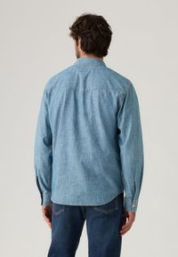 Light blue denim shirt with long sleeves, button collar, and back yoke detail. Features a straight hem and buttoned cuffs.